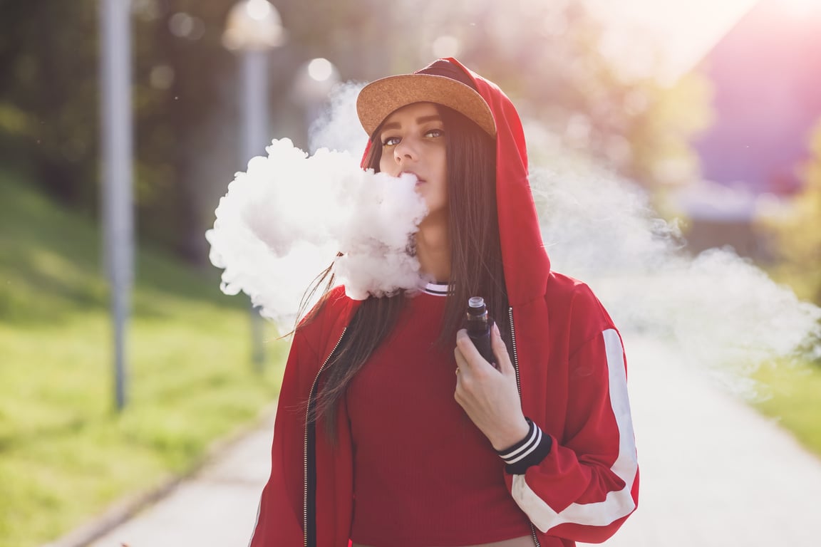 Vaping Girl. Young Woman Vape E-Cig. Pretty Young Female in Black Hat, Red Clothing Vape Ecig, Vaping Device at the Sunset. Toned Image. Hip-Hop Style.